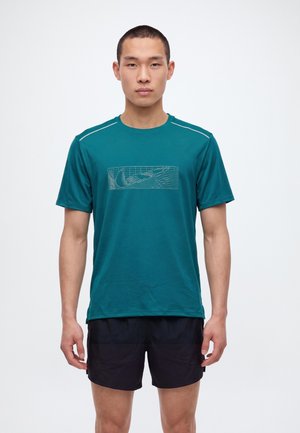 Un homme se tient face à l’objectif, vêtu d’un tee-shirt de sport bleu canard avec un motif sur la poitrine et d’un short de sport noir, sur un fond blanc uni.