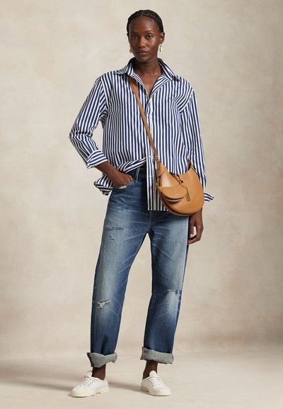 Maglietta a righe blu e bianche, jeans chiari strappati, borsa a tracolla marrone e sneakers bianche. Il modello è in piedi con le mani in tasca.