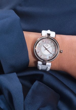 White silicone strap watch with a round silver-tone case, white dial, Roman numerals, and crystal accents. Worn on a wrist over dark fabric.