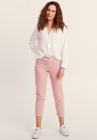 Weißes langärmliges Blusenshirt mit Knopfdetails, kombiniert mit hellrosa, kurzen Hosen mit elastischem Bund und umgeschlagenen Bündchen, getragen mit weißen Sneakers.