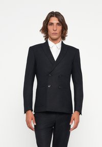 Blazer de corte cruzado en un elegante azul marino, con tejido texturizado, solapas amplias, dos bolsillos frontales y sutiles detalles de botones. Se ven pantalones a juego.