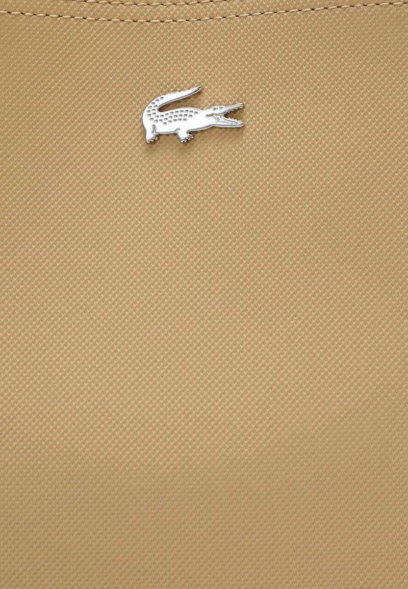 Tela texturizada en color beige con un patrón sutil, que presenta un acento de logo de cocodrilo plateado en la esquina superior izquierda.