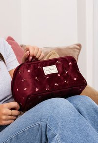 Bordowy welurowy worek kosmetyczny z haftem kwiatowym, prostokątny kształt, z zamkiem błyskawicznym i metką markową. Miękka tekstura i szczegółowy design.