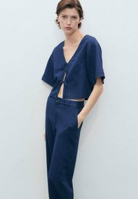 Temno moder skrajšan top z kratkimi rokavi in sprednjim zavezovanjem, v kombinaciji z visokim pasom in ravnimi hlačami v temno modri barvi, ki imajo gladko teksturo.