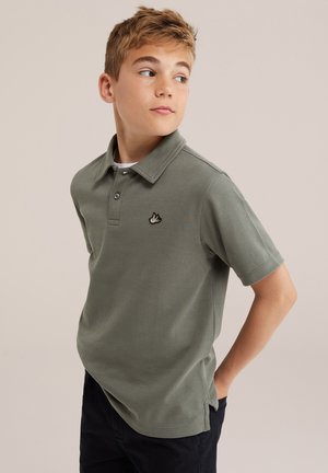 Garçon aux cheveux courts châtain clair portant un polo vert avec un logo d'oiseau, regardant vers la droite avec une main dans la poche, sur un fond uni.