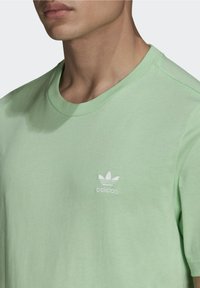 Světle zelené tričko z jemné bavlny s kulatým výstřihem. Na levé části hrudi se nachází bílé logo adidas a tričko má krátké rukávy.