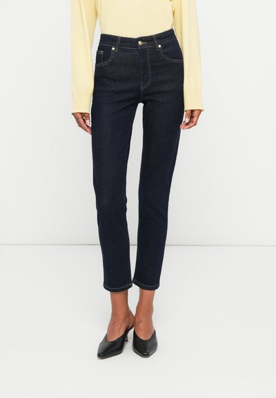 Jean en denim bleu foncé avec une taille haute, coupe slim et revers. Présente des accents de boutons et de coutures dorés. Associé à des mocassins noirs à talons pointus.