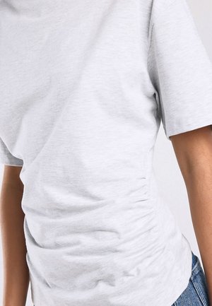 Close-up van een persoon die een lichtgrijs shirt met korte mouwen en een blauwe spijkerbroek draagt, waarbij de torso en de details van de arm zichtbaar zijn.