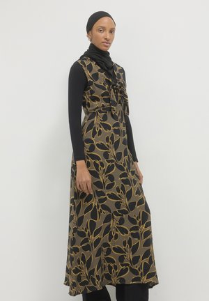 Maxi dress in black and gold floral pattern, with a tied neckline. Made of textured fabric, layered over a long-sleeve top and pant.