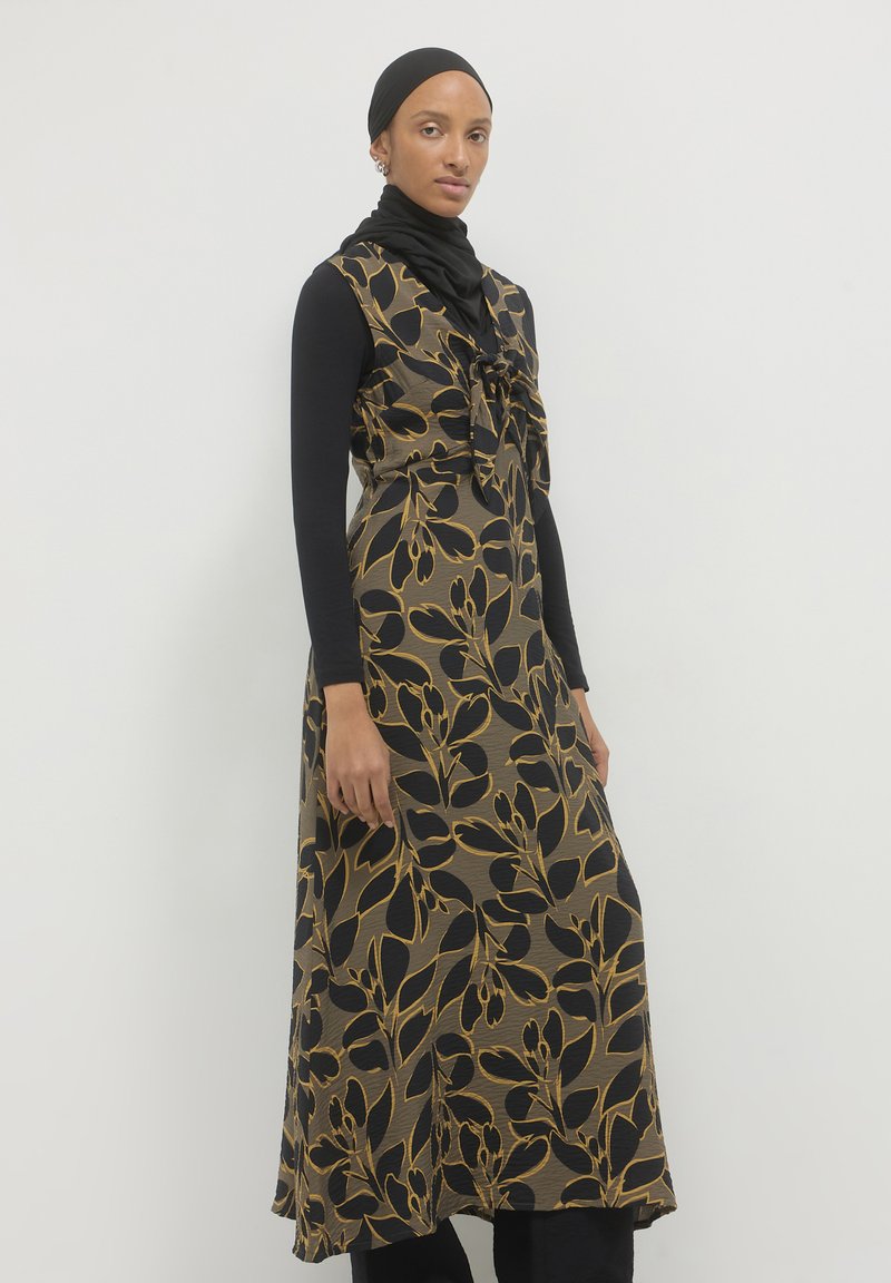 Maxikleid in schwarz mit goldfarbenem Blumenmuster und gebundenem Ausschnitt. Hergestellt aus strukturiertem Stoff, über einem Langarmoberteil und einer Hose geschichtet.