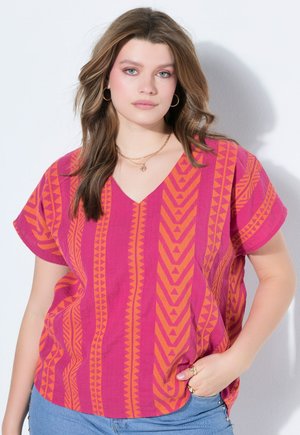 Ung kvinde med bølget hår bærer hoop-øreringe, lagdelt guldkæder, en pink og orange mønstret V-hals bluse og blå jeans med nitte detaljer.