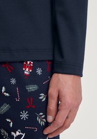 Marineblauw shirt met lange mouwen, gecombineerd met een marineblauw onderstuk met patronen van feestelijke motieven zoals zuurstokken, sneeuwvlokken en hulstbladeren.