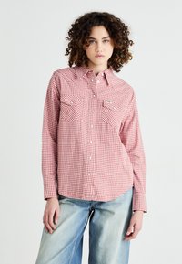 Chemise à carreaux rouges et blancs avec manches longues, patte de boutonnage, deux poches poitrine et ourlet arrondi, fabriquée à partir d'un tissu léger.