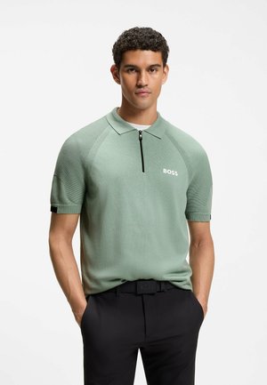 Young man with short curly hair wearing a light green short-sleeve polo with a black zipper and black pants, hands in pockets.