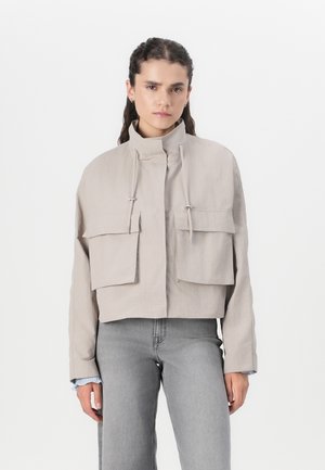Beige Jacke mit cropped Schnitt, hohem Kragen, zwei Fronttaschen, Kordelzügen und glatter Textur, kombiniert mit grauen Jeans.