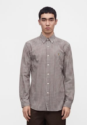 Jeune homme portant une chemise à manches longues à carreaux marron et blanc avec un col pointu, debout devant un fond blanc uni.