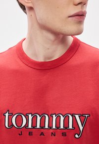 Camiseta de algodón roja con cuello redondo, bordada con "tommy" sobre "JEANS" en colores contrastantes, mostrando una textura suave.