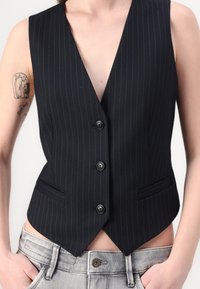 Schwarze, gestreifte Weste mit tiefem V-Ausschnitt, fünf Knöpfen, Seitentaschen und taillierter Silhouette, kombiniert mit hellen Jeans.