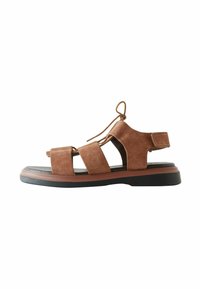 Sandal de ante marrón con múltiples correas, lados abiertos, un cierre atado y una suela de goma plana con un borde contrastante.