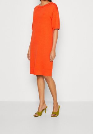 Vrouw draagt een oranje jurk tot op de knie met korte mouwen, gecombineerd met olijfgroene, open-toe hakmuilen, staand tegen een witte achtergrond.