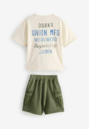 T-shirt crème avec un texte bleu et marron affichant divers noms de marques, associé à un short cargo vert olive avec des poches latérales.