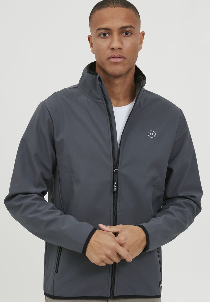Charcoal gray softshell jacket with a high collar, zip closure, two side pockets, and elastic cuffs. Reinforced stitching and logo detail.