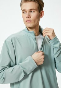 Chaqueta de cremallera verde claro con un alto cuello, mangas largas y una textura suave. Presenta una camiseta blanca parcialmente visible debajo.