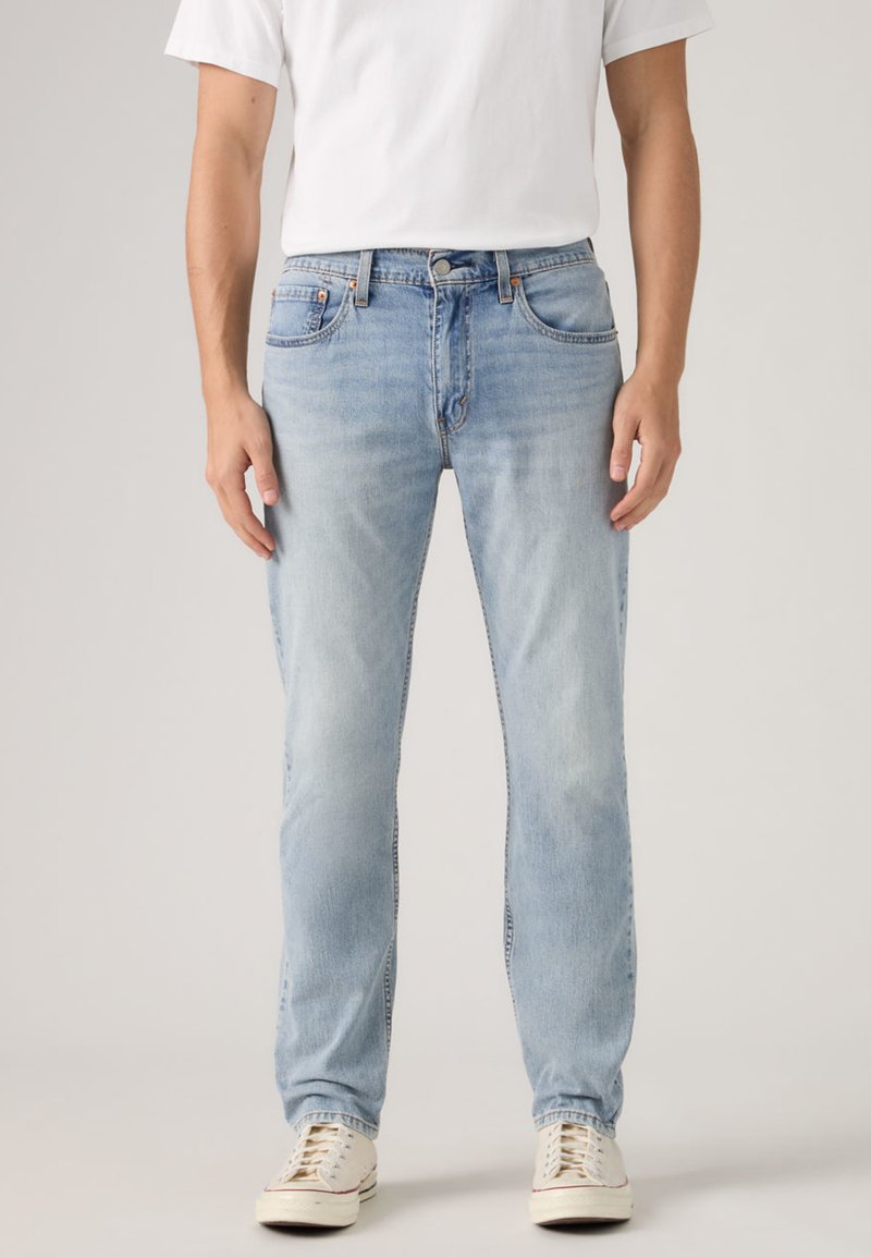 Άνδρας που φοράει τζιν σε ίσια γραμμή ανοιχτού μπλε χρώματος, λευκό μπλουζάκι και λευκά αθλητικά παπούτσια, στέκεται μπροστά σε απλό φόντο.