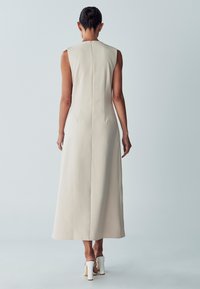 Sleeveless, ankle-length dress in light beige fabric, featuring a concealed back zipper and a tailored fit. Worn with white high-heeled shoes.