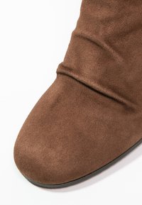 Braune Wildlederstiefel mit abgerundetem Zeh und gefalteten Details an der Oberseite, vor einem weißen Hintergrund.