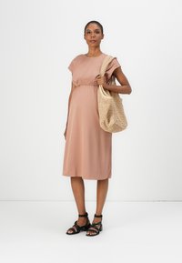 Maternity dress in soft blush fabric with short sleeves and gathered waist. Paired with a woven beige bag and black strappy sandals.