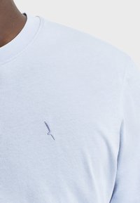 Maglietta a maniche corte di colore azzurro chiaro con colletto rotondo. Presenta un piccolo logo ricamato in una tonalità leggermente più scura sul lato sinistro del petto.