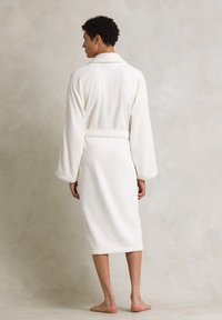 Accappatoio bianco in spugna con maniche lunghe, colletto a scialle e vita legata. Texture morbida e soffice che arriva fino al ginocchio. Posizione a piedi nudi.