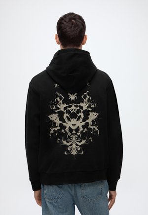 Homme portant un sweat à capuche noir avec un motif floral symétrique beige dans le dos, associé à un jean bleu, tourné dos à un fond uni.