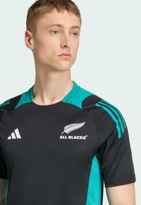 Jersey atlético negro con acentos en color teal. Presenta un cuello redondo, mangas cortas, un logotipo blanco de "ALL BLACKS" y tres franjas blancas en los hombros.