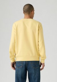Man met een kaal hoofd in een effen gele sweatshirt en blauwe spijkerbroek, staand met zijn rug naar de camera tegen een neutrale achtergrond.