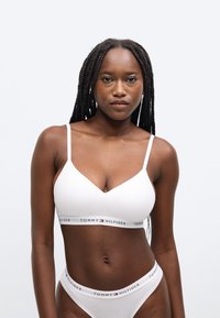 Biela bavlnená braletka s výstrihom do V a tenkými ramienkami. Má elastickú pásku s logom "TOMMY HILFIGER" v červenej a modrej farbe.