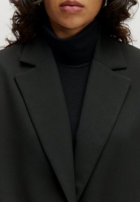 Zwarte blazer met een inkeping op de revers en gestructureerde schouders, gedragen over een zwarte geribde coltrui. Gladde textuur, minimalistisch ontwerp.