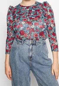 Blomstret bluse med volanger på ærmerne, der har en blanding af røde, blå og grønne blomster på et gennemsigtigt stof. Sat sammen med højtaljede denimjeans.