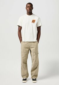 Camiseta de color crema con el logo "Casey Jones" en naranja, combinada con pantalones cargo beige que presentan bolsillos y detalles de costura. Zapatillas en blanco y negro.