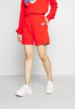 Personne portant un short à taille élastique rouge et un sweat assorti, avec des baskets blanches épaisses, debout devant un fond uni.