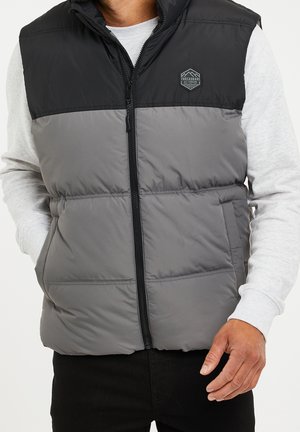 Homme portant une veste matelassée noire et grise avec le logo "Threadbare All Terrain", une chemise blanche à manches longues et un pantalon noir, mains détendues.