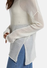 Leichtes, cremefarbenes Strickpullover mit vertikalen Streifen, seitlichen Schlitzen und verlängerten Ärmeln. Er bietet eine lockere Passform und eine strukturierte Textur.