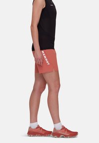 Różowe szorty z białym logo "MAMMUT" po lewej stronie, zestawione z czarnym bezrękawnikem i koralowymi butami do biegania z jasnymi akcentami.