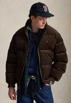 Junger Mann, der eine dunkelbraune Daunenjacke, blaue Jeans, einen karierten Schal, einen blauen Pullover und eine dunkle Baseballkappe trägt und seitlich nach unten schaut.