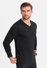 Maglietta nera a maniche lunghe e scollo a V, realizzata in materiale morbido, design aderente con delicate cuciture laterali, abbinata a jeans scuri e un orologio.