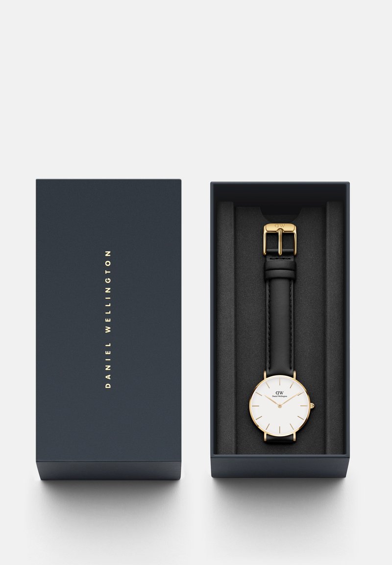 Schwarze Lederarmbanduhr mit weißem Zifferblatt, goldenen Akzenten und minimalistischen Design, präsentiert in einer marineblauen Box mit schwarzem Innenraum.