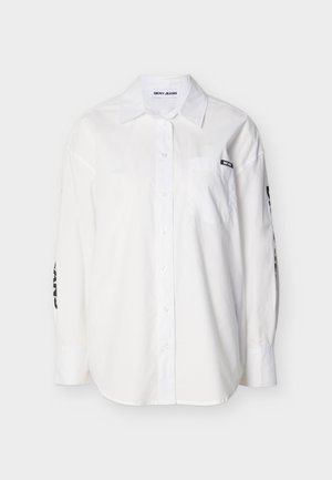 Hvit langermet skjorte med knapper, krage, frontlomme og svart "DKNY JEANS"-etikett på lommen og ermene.