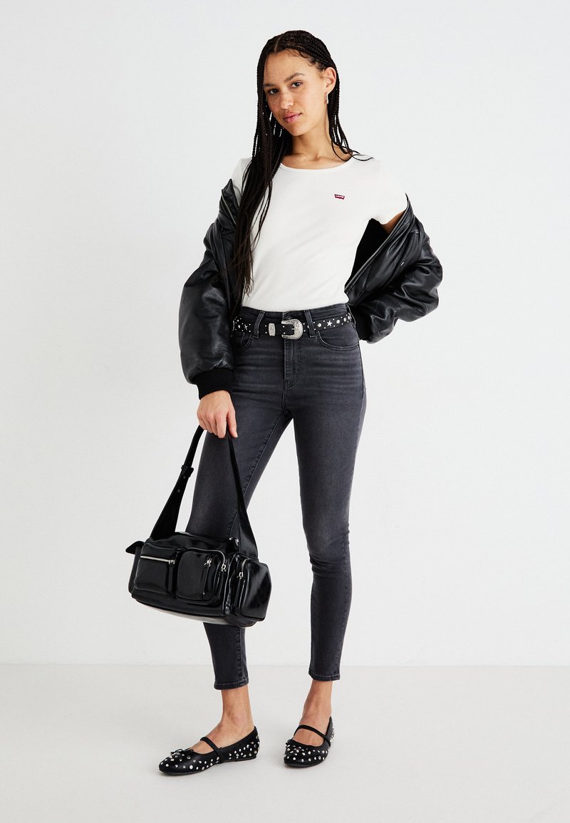 Weißes T-Shirt, schwarze oversized Bomberjacke, dunkle Skinny-Jeans, besetzte schwarze Ballerinas und eine schwarze Handtasche mit mehreren Reißverschlusstaschen.
