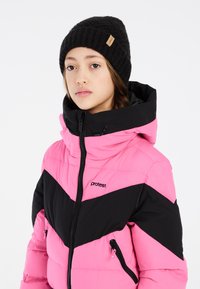 Doudoune rose et noire avec un design en chevrons, dotée d'un col montant, de fermetures éclair et du logo de la marque sur la poitrine. Bonnet en tricot noir.